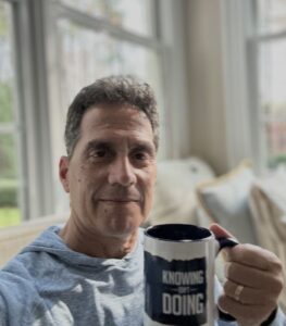 A man in a light blue hooded sweatshirt is holding a white mug with a dark blue bottom band. The mug has the phrase "KNOWING ISN'T DOING" printed on it in white text. He is seated indoors near large windows that let in bright light.