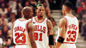 Jordan, Dennis Rodman, and Scottie Pippen of the 1990s Chicago Bulls standing together on the court in their iconic red and white home jerseys.