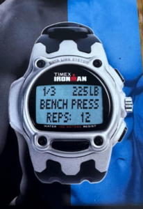 A white and black Timex Ironman digital watch displaying workout tracking data on its screen, reading "1/3 225LB BENCH PRESS REPS: 12".